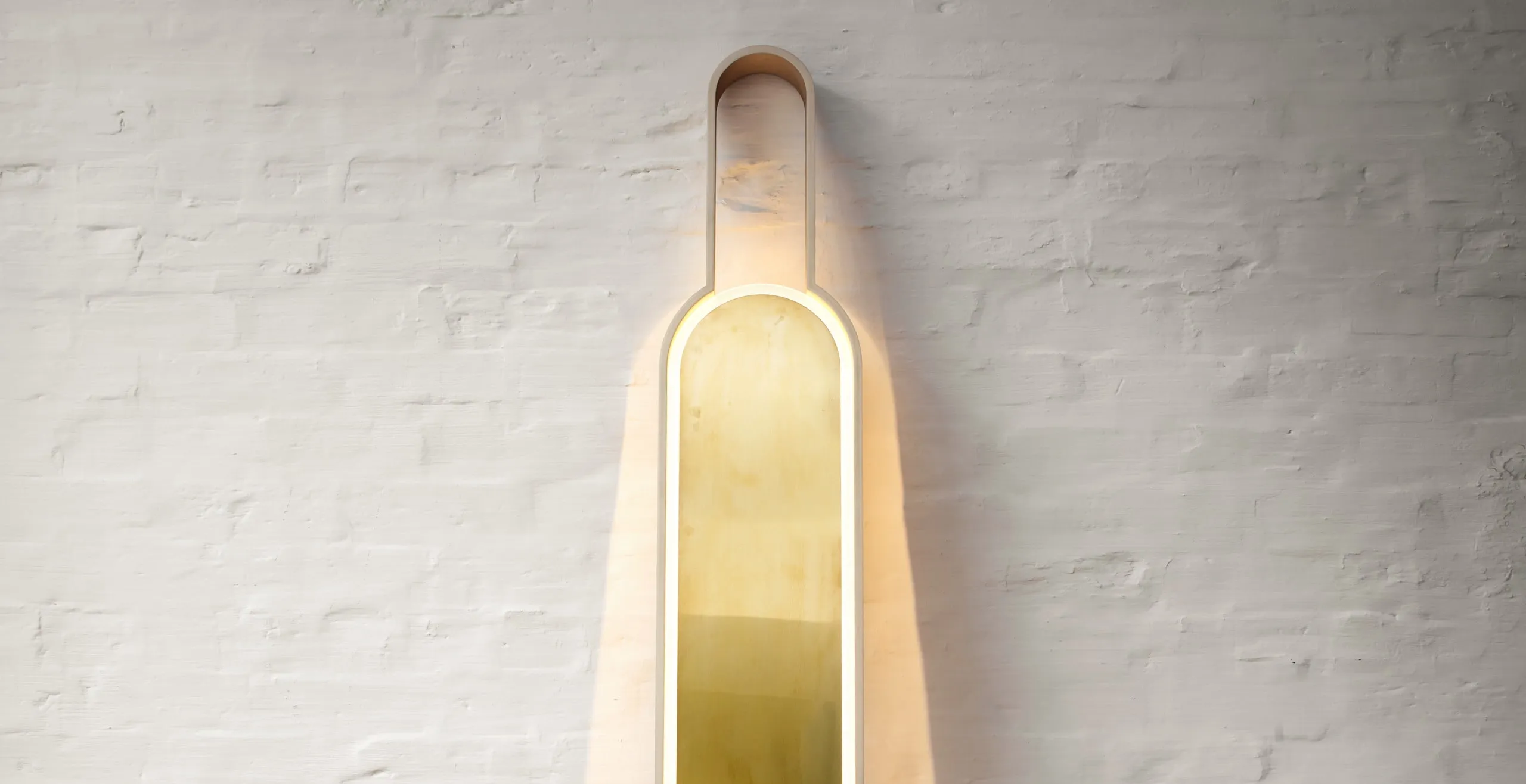 Portal Floor Lamp in brass, lit, leaning against a white brick wall