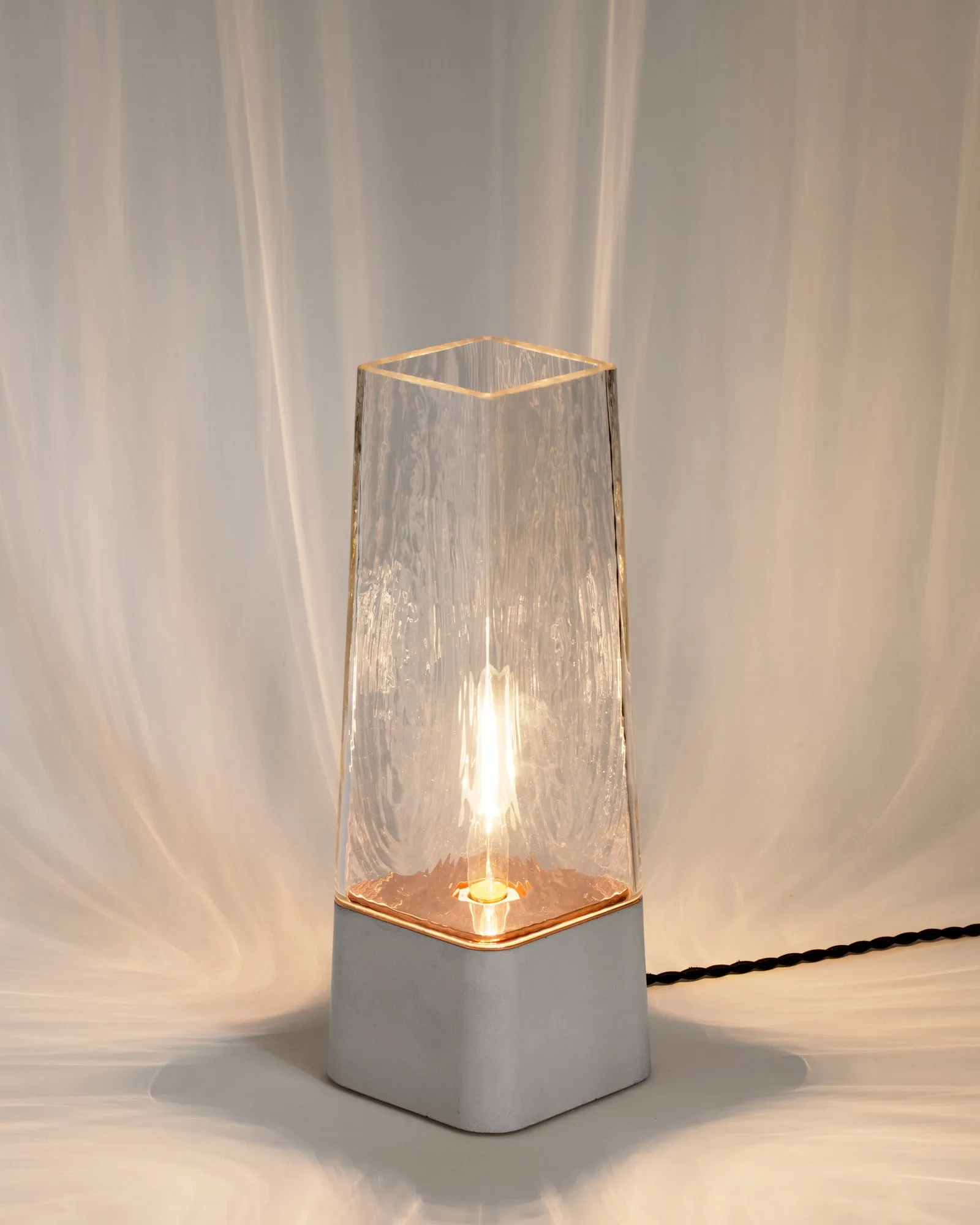 Faaro in clear glass on a white concrete base, lit, before a pale drape