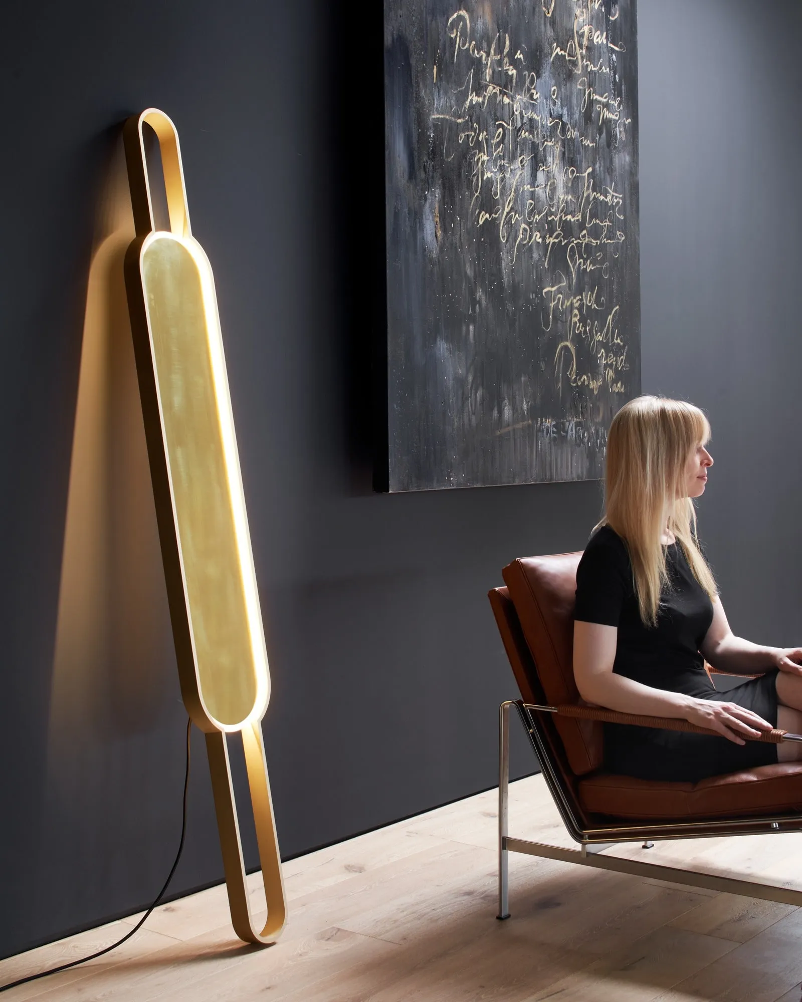 Portal Floor Lamp lit beside a woman in a leather armchair, a dark painting on the wall