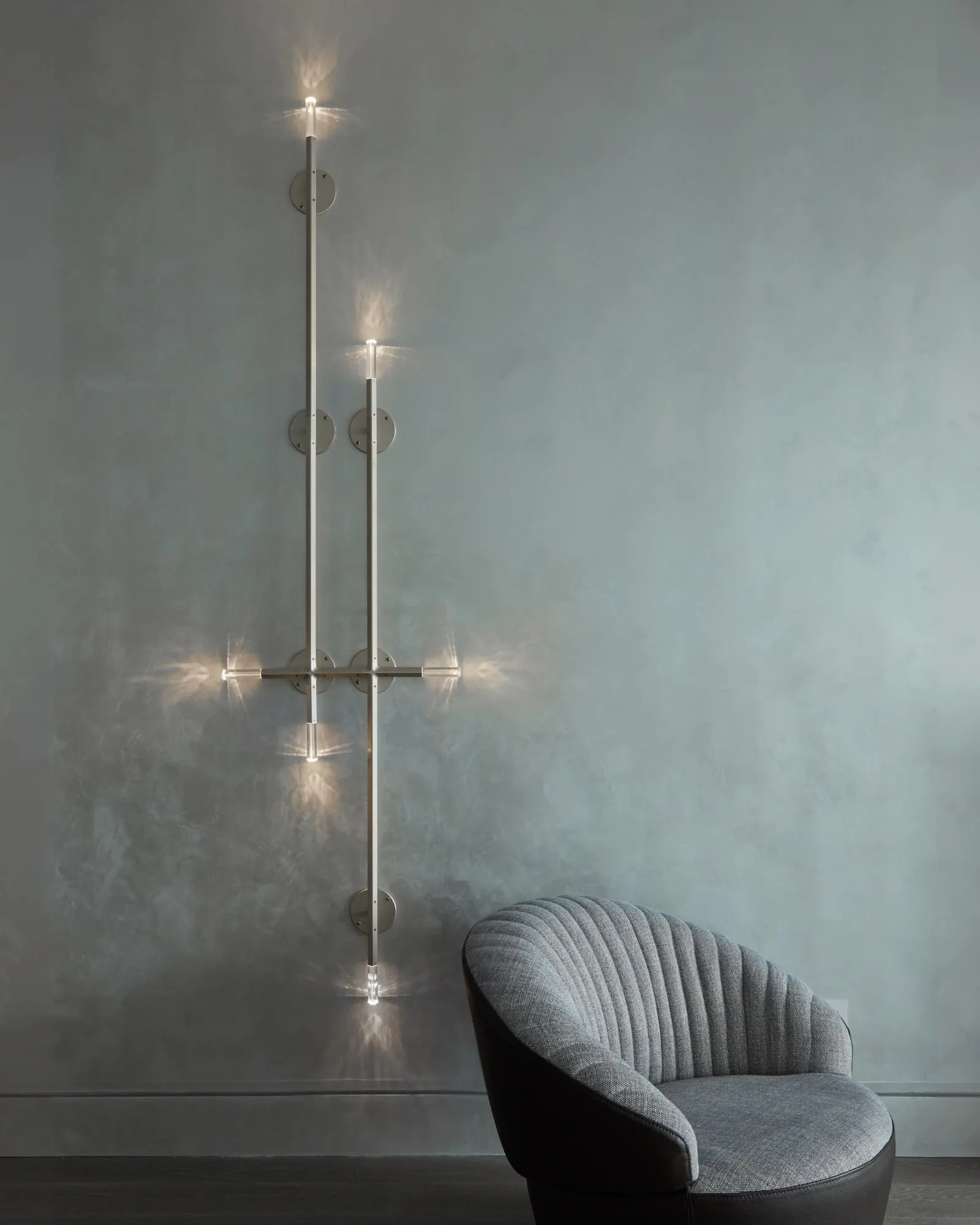 A tall JAX wall sconce in Satin Brass beside a grey armchair