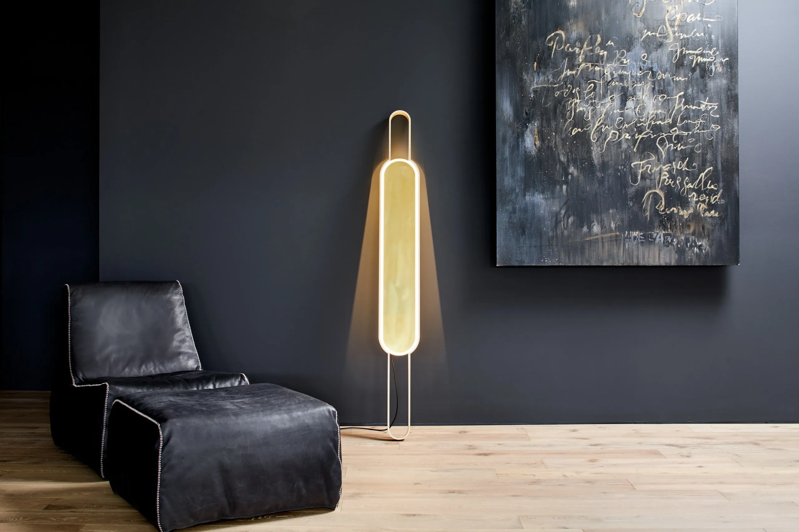 Portal Floor Lamp lit against a charcoal wall beside a leather chaise and an abstract painting