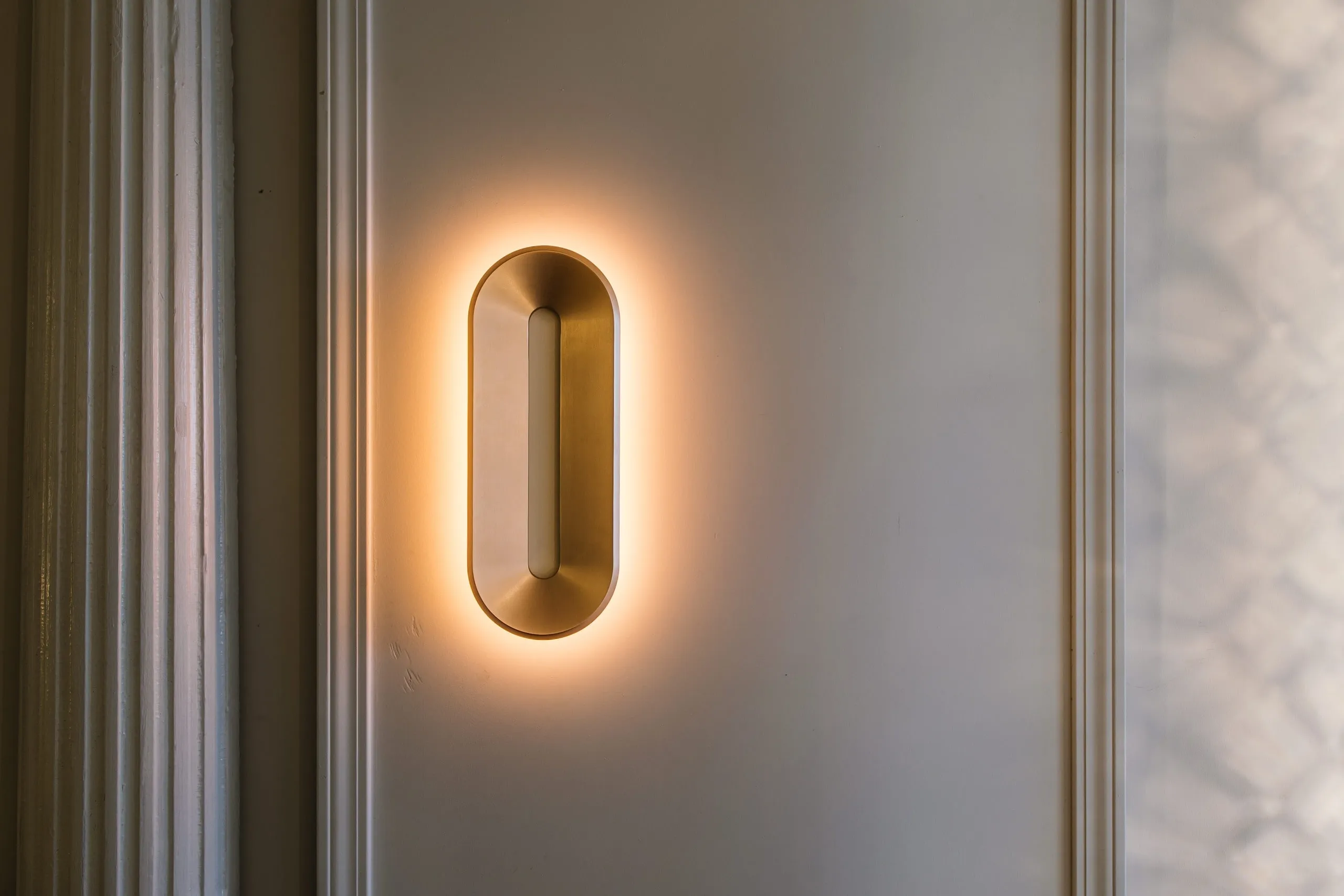 Void Sconce in brass casting a ring of light on a pale wall between moldings