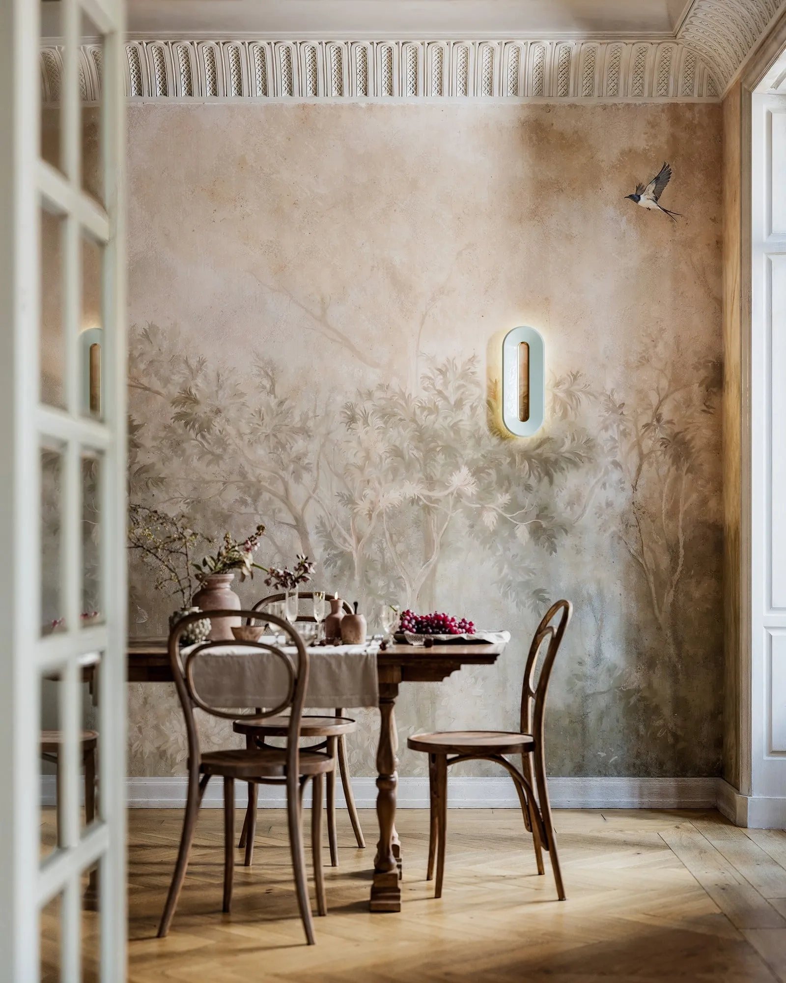 Void Ceramic Sconce above a dining table in a room with a painted mural, off, then lit as it reaches the middle of the screen